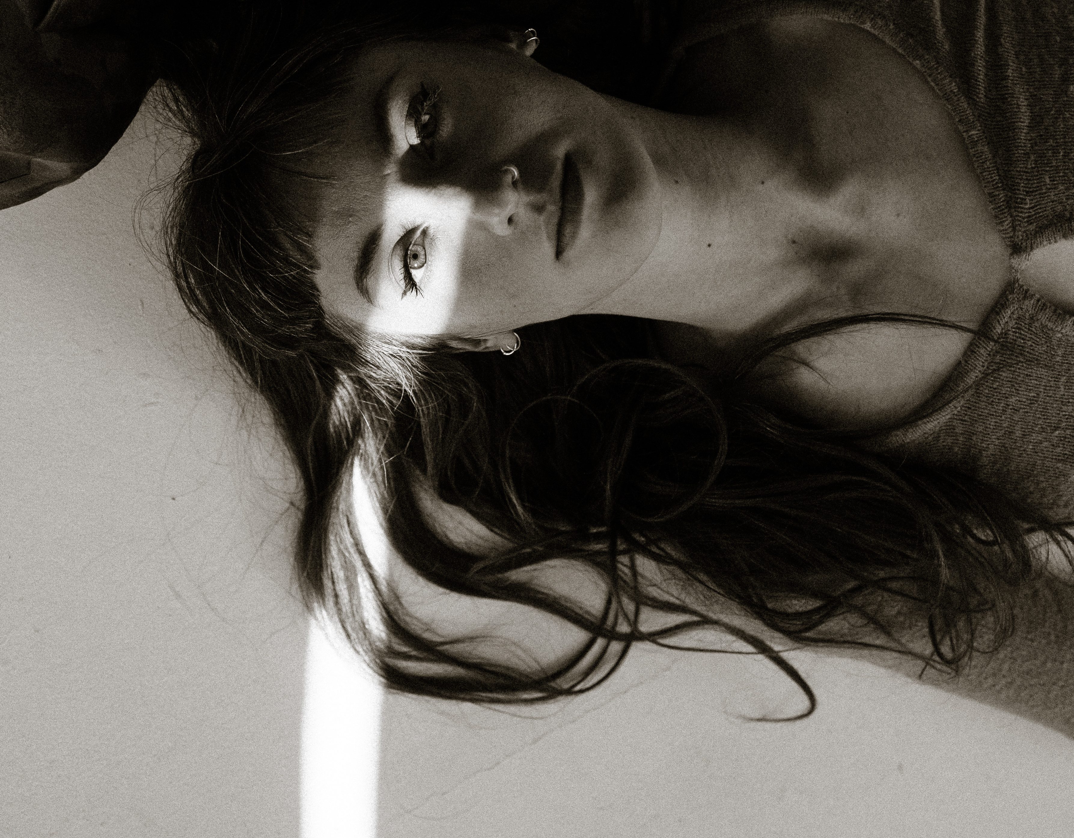 a black and white image of Charlotte, laying on the floor with her hair spread around her. She has dark long hair with a fringe, and a sunbeam is shining over her eyes.